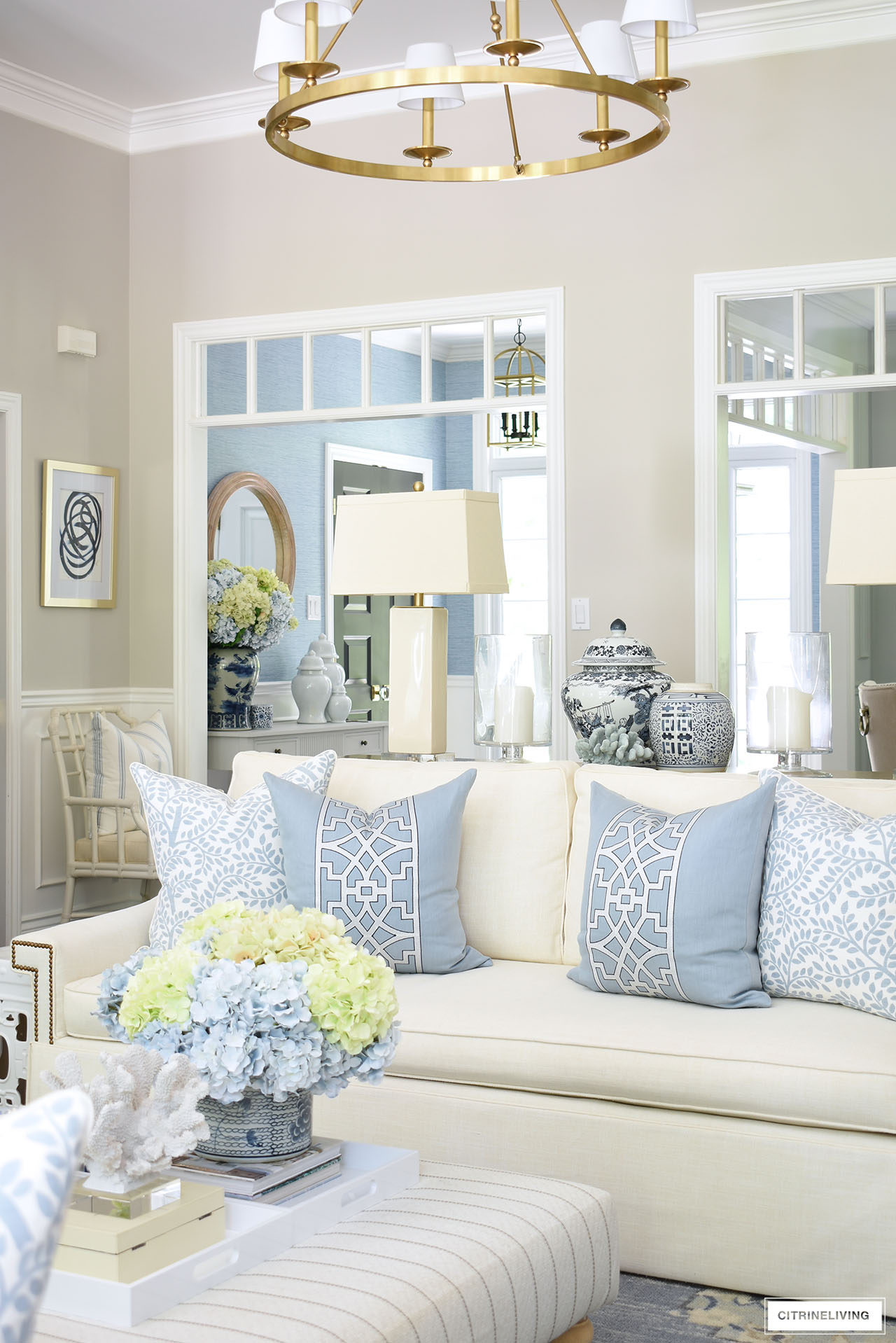 Elegant blue and white pillows styled on a white sofa, a light green and blue hydrangea arrangement sits in front on an upholstered ottoman. Blue and white ginger jars styled throughout the room and beyond.