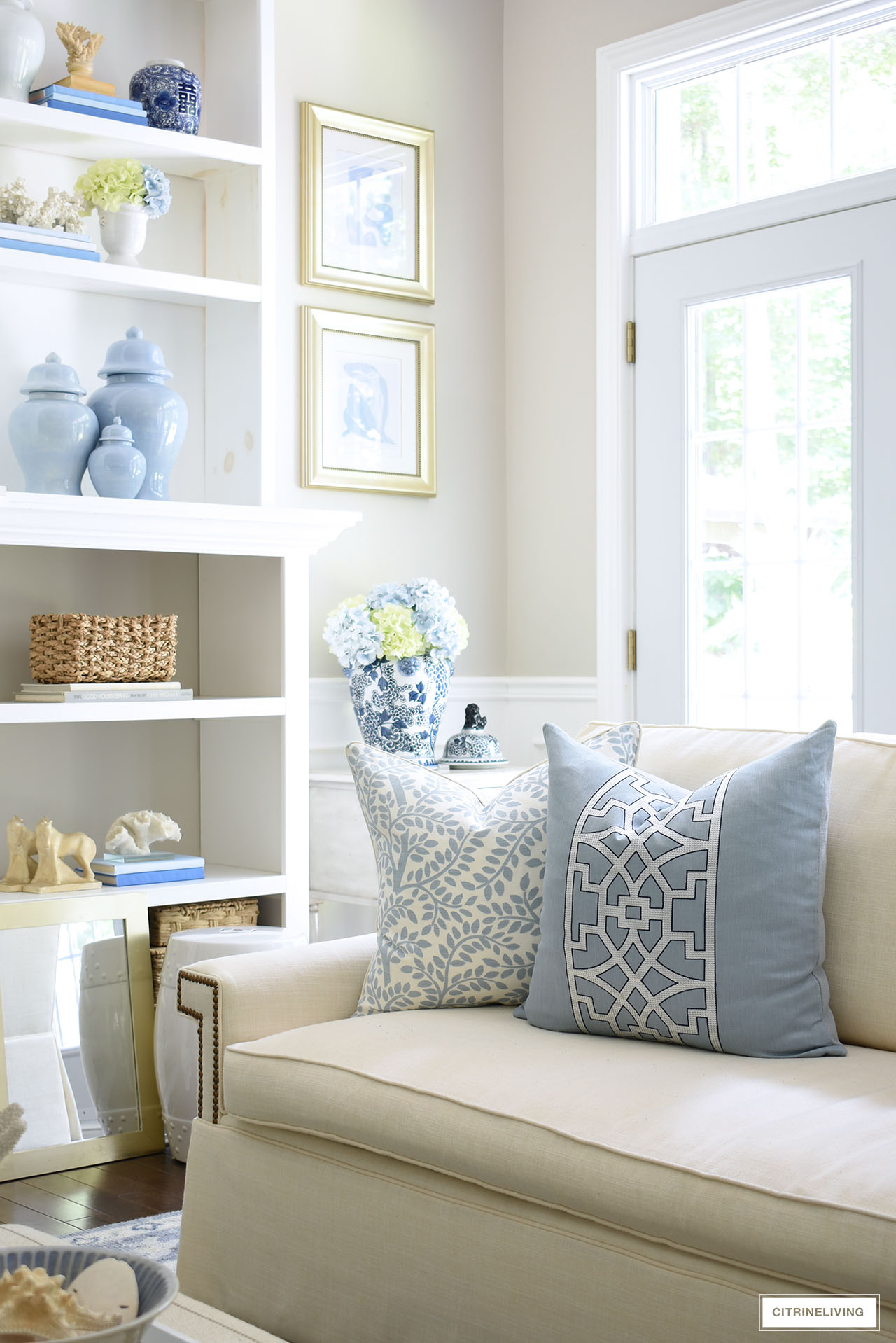 A living room corner styled for summer with faux hydrangeas, blue and white accents and designer pillows on a white sofa.