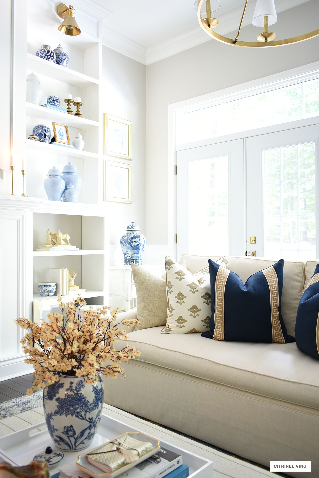 Gorgeous designer throw pillows in navy blue, tan and ivory, styled for fall in an elegant living room, with faux flowers and gold candlesticks.
