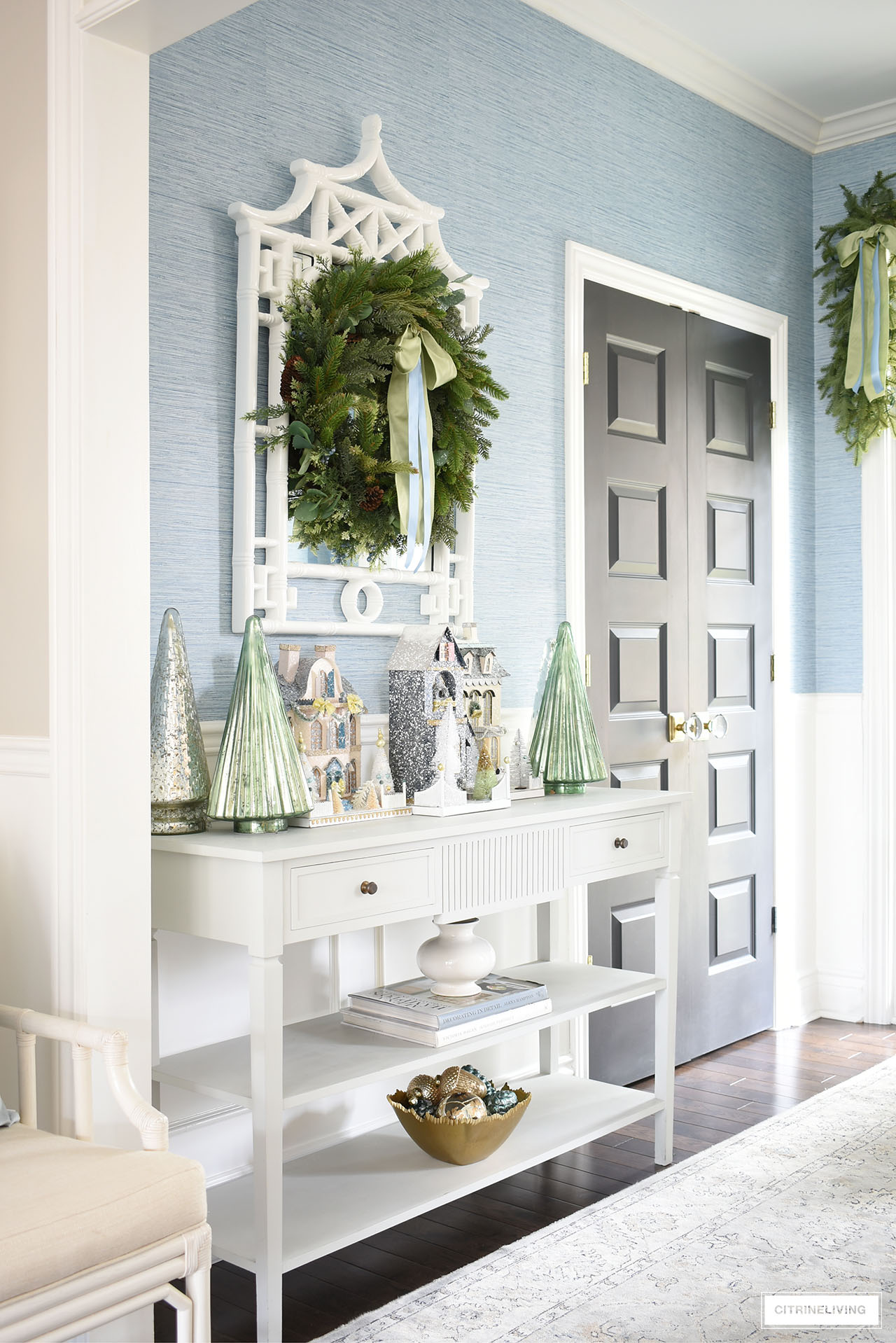 Christmas entryway decorated with Christmas houses, mercury glass Christmas trees and a lush green wreath decorated with a beautiful light green and light blue bow.