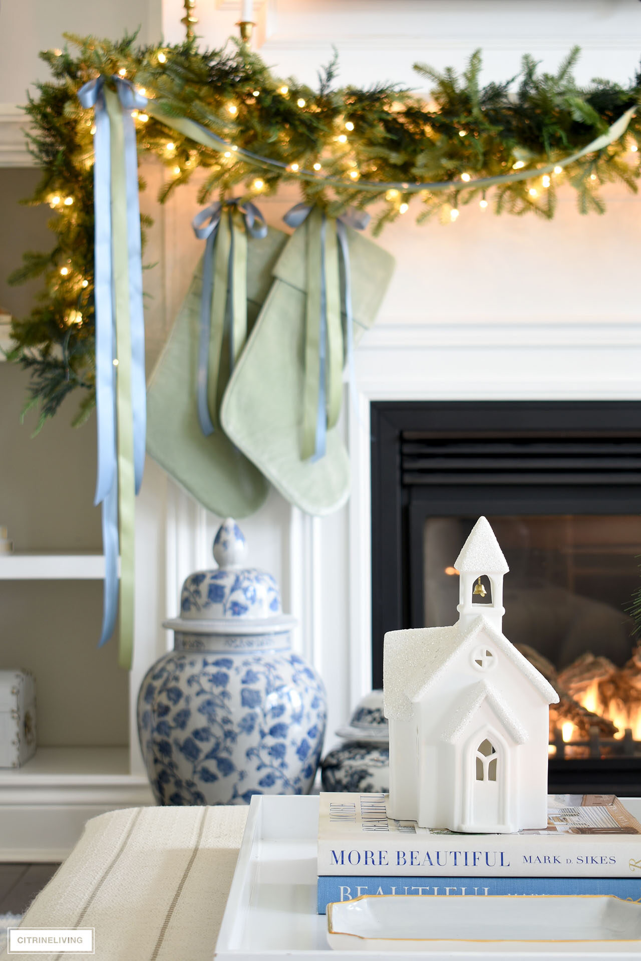 Blue and green Christmas decor, coffee table styling with a white ceramic church, and Christmas mantel with green stockings in the background.