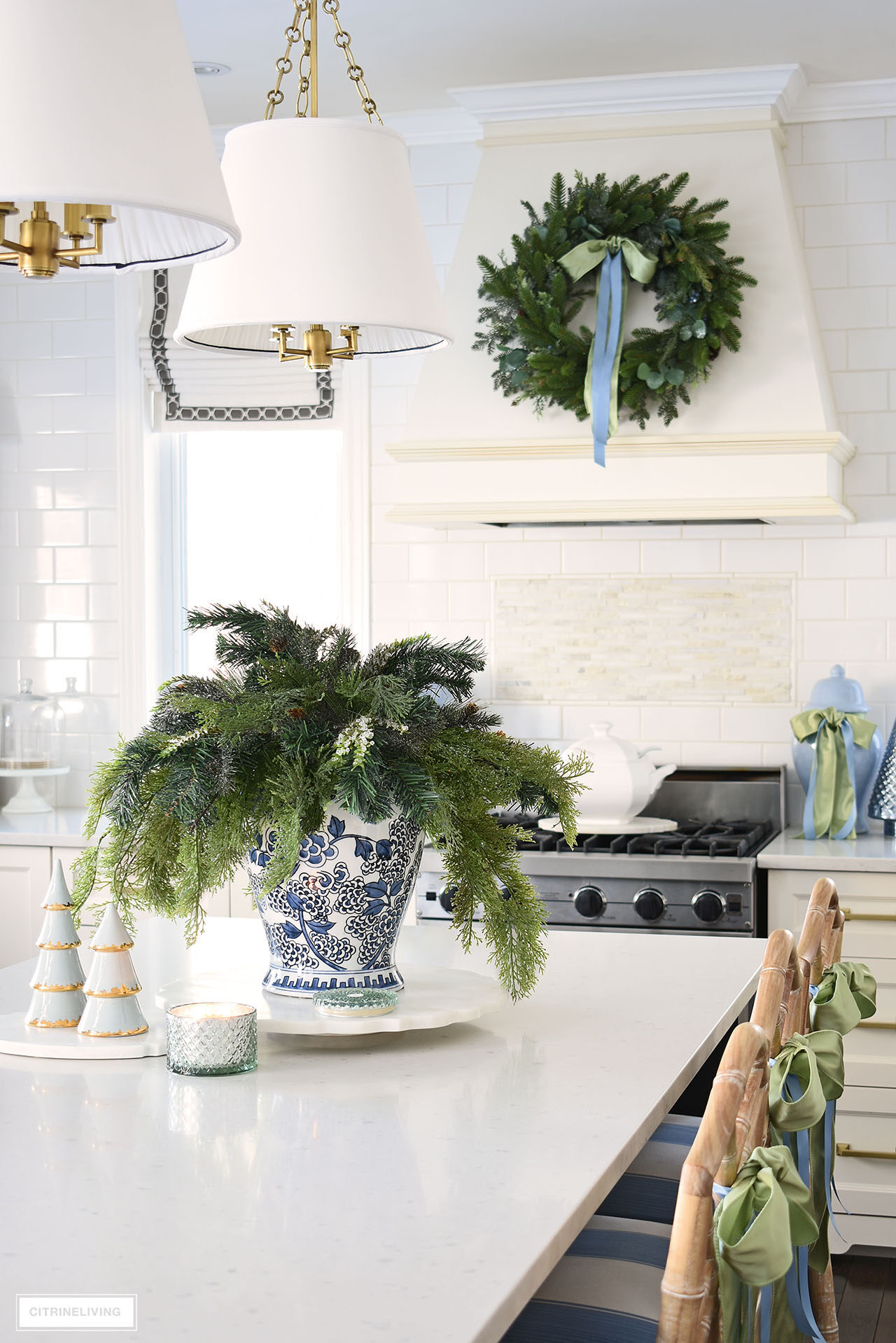 A Christmas vignette on a kitchen island with blue and white ginger jar and greenery arrangement. A beautiful green wreath hangs on the range hood in the background.