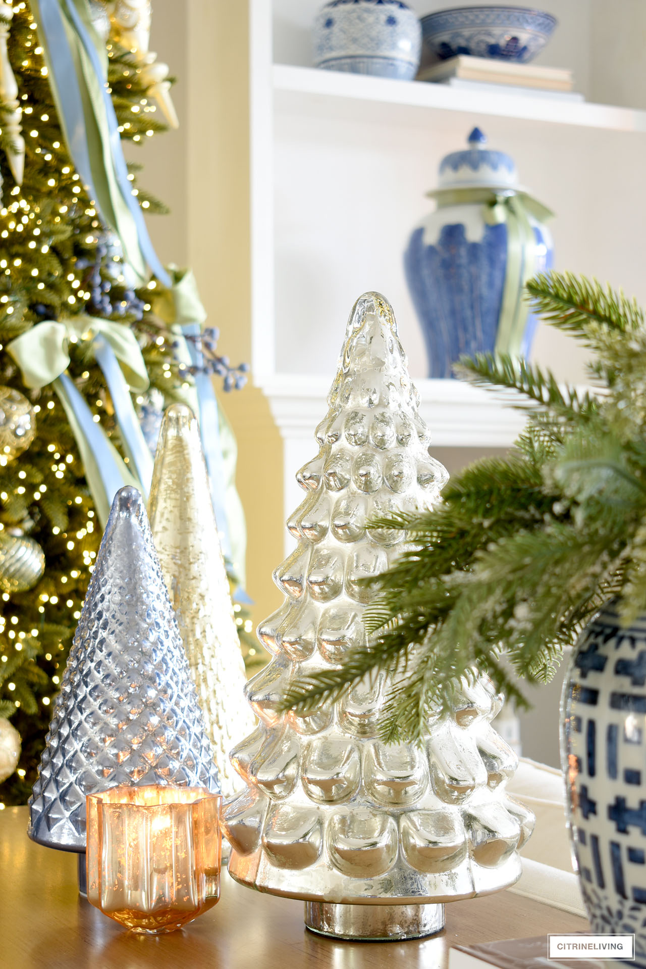 Mercury glass Christmas trees in silver, blue, gold and green with a greenery arrangement in a blue and white ginger jar.