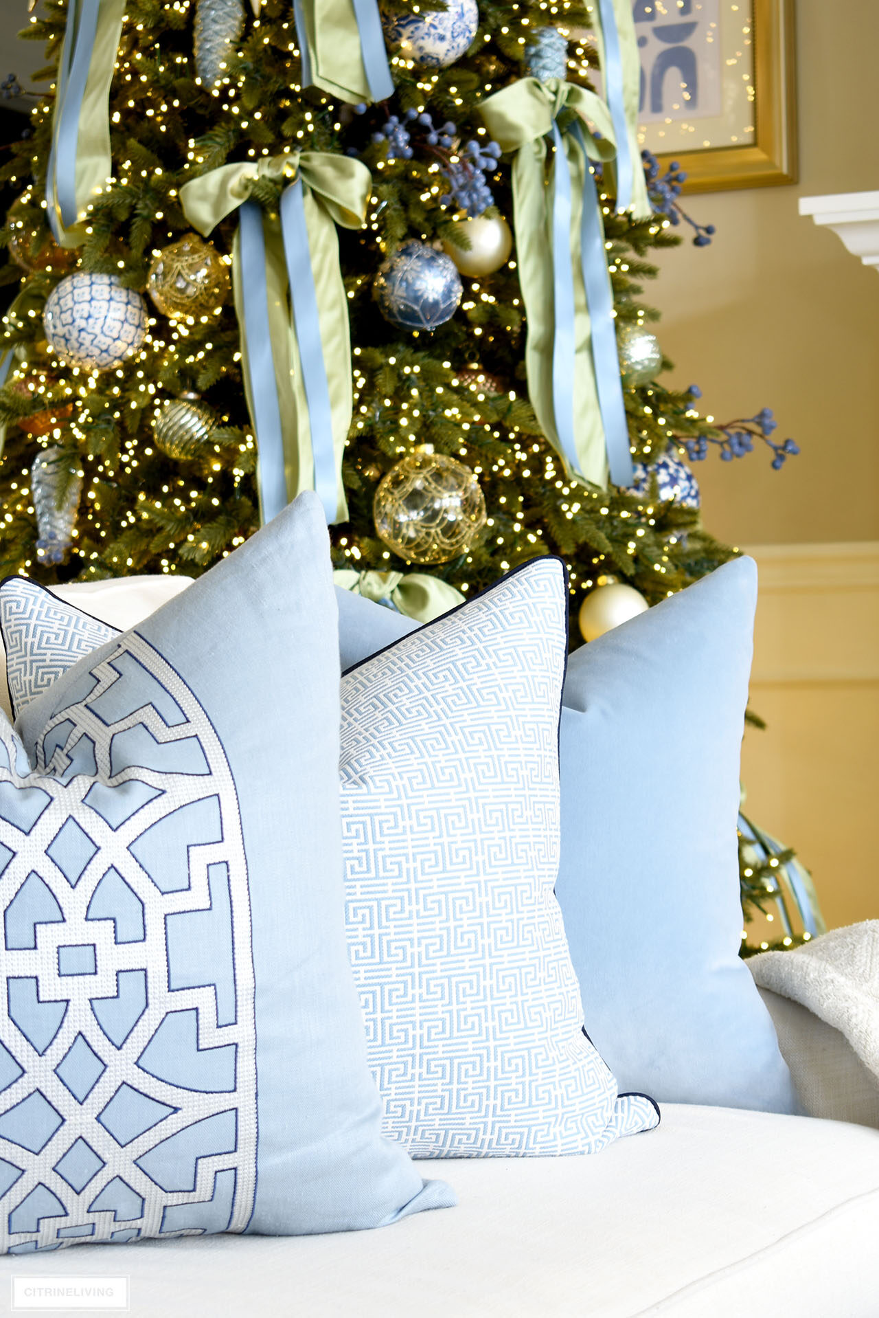 Blue and white throw pillows on a white sofa, Christmas tree decorated with light green and blue bows sits behind.