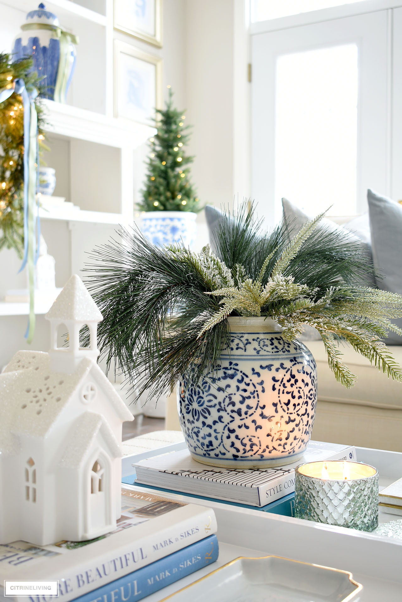 A holiday arrangement with Christmas greenery in a blue and white ginger jar styled with a light green scented candle, white Christmas village church.