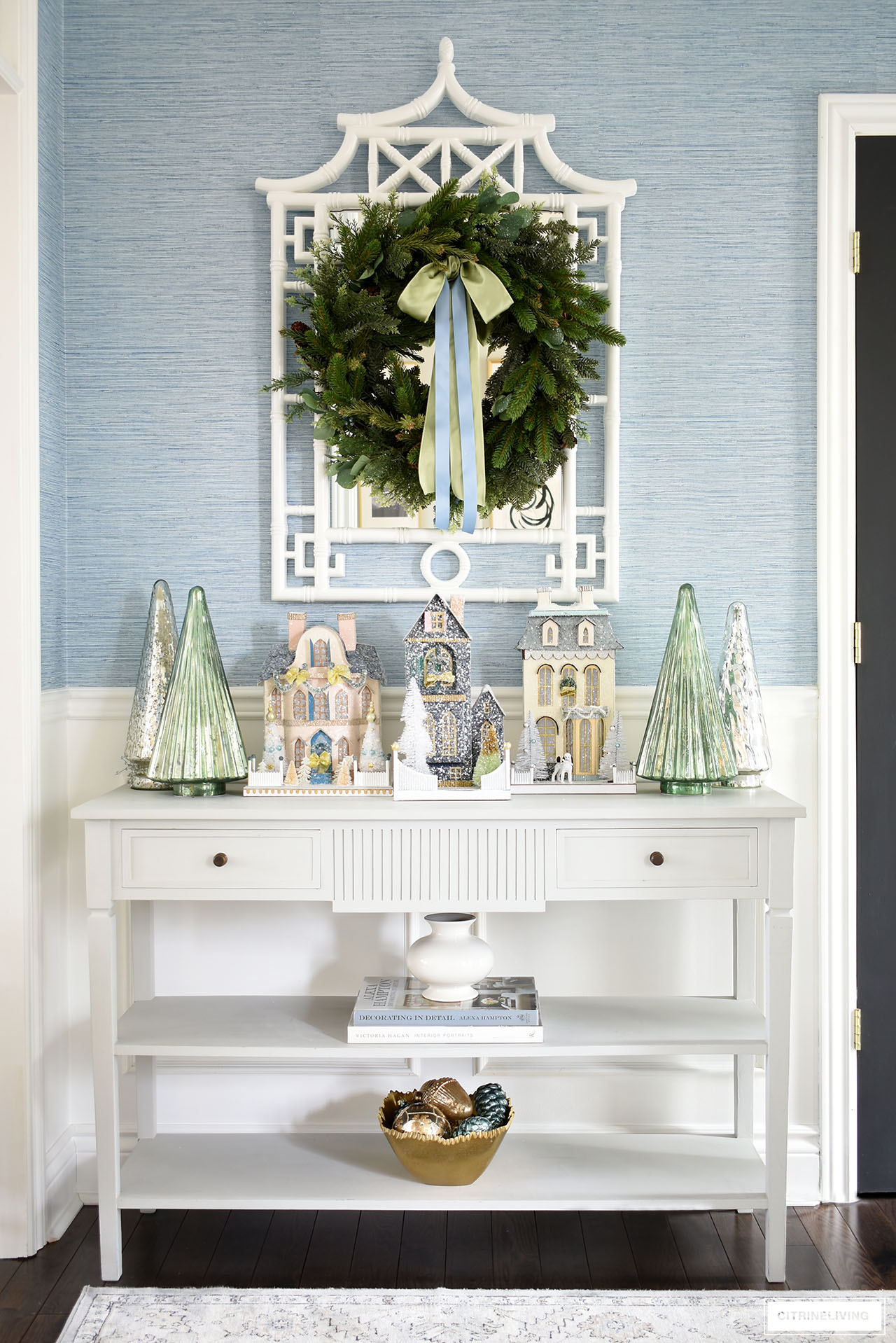A gorgeous Christmas display on an entryway console table styled with vintage inspired Christmas houses, mercury glass trees and a lush green wreath hanging about on a white pagoda mirror.