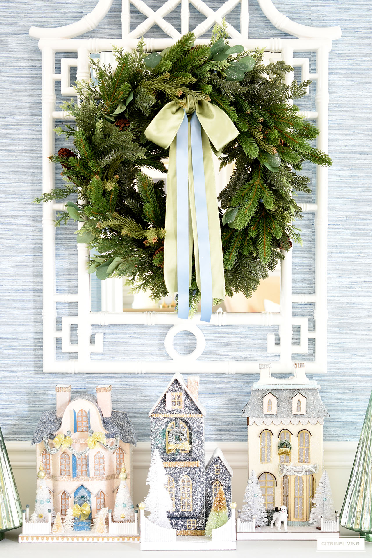 A stunning Christmas display on an entryway console table using vintage inspired Christmas houses, and a lush green wreath hanging about on a white pagoda mirror.