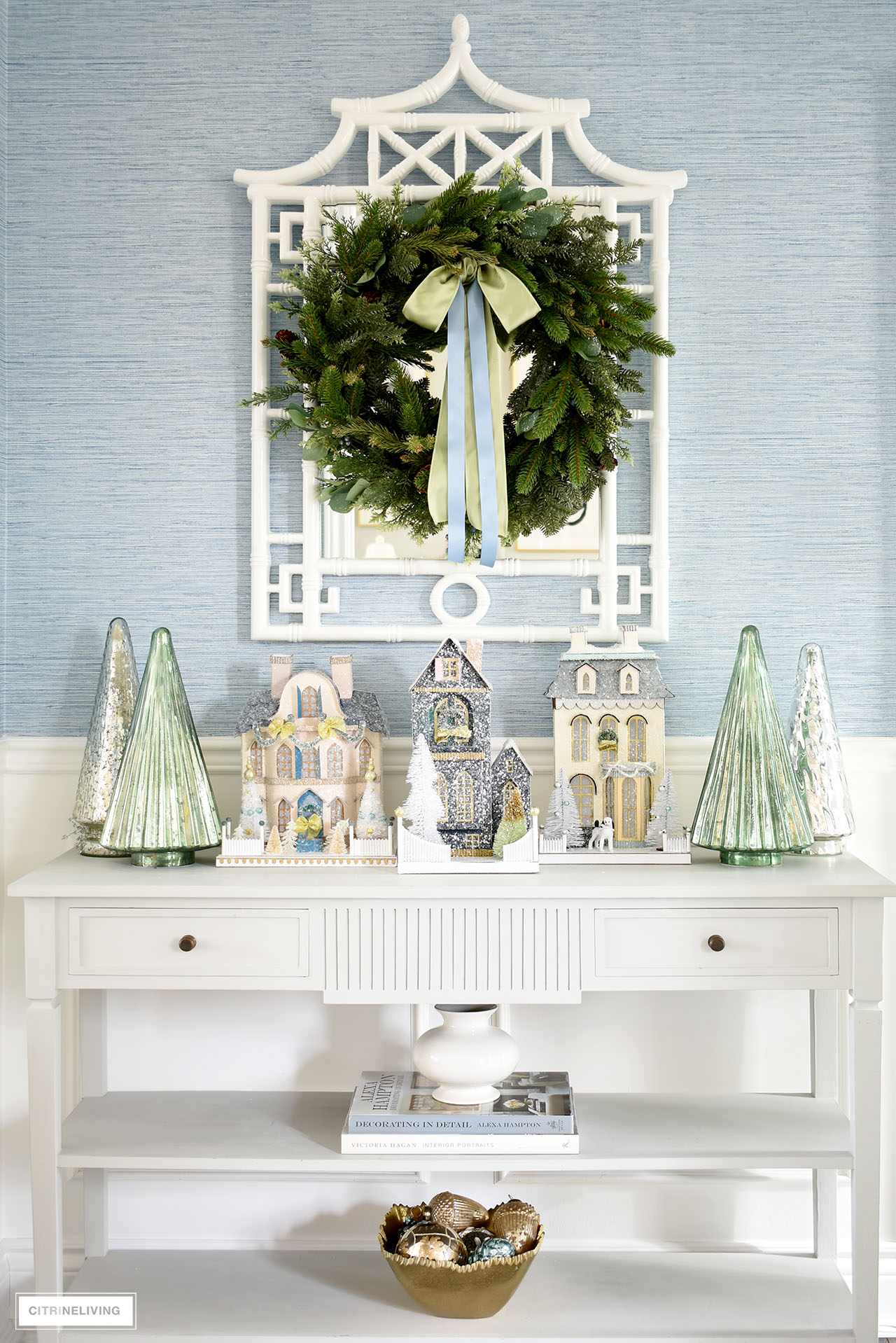 A stunning Christmas display on an entryway console table using vintage inspired Christmas houses, mercury glass trees and a lush green wreath hanging about on a white pagoda mirror.