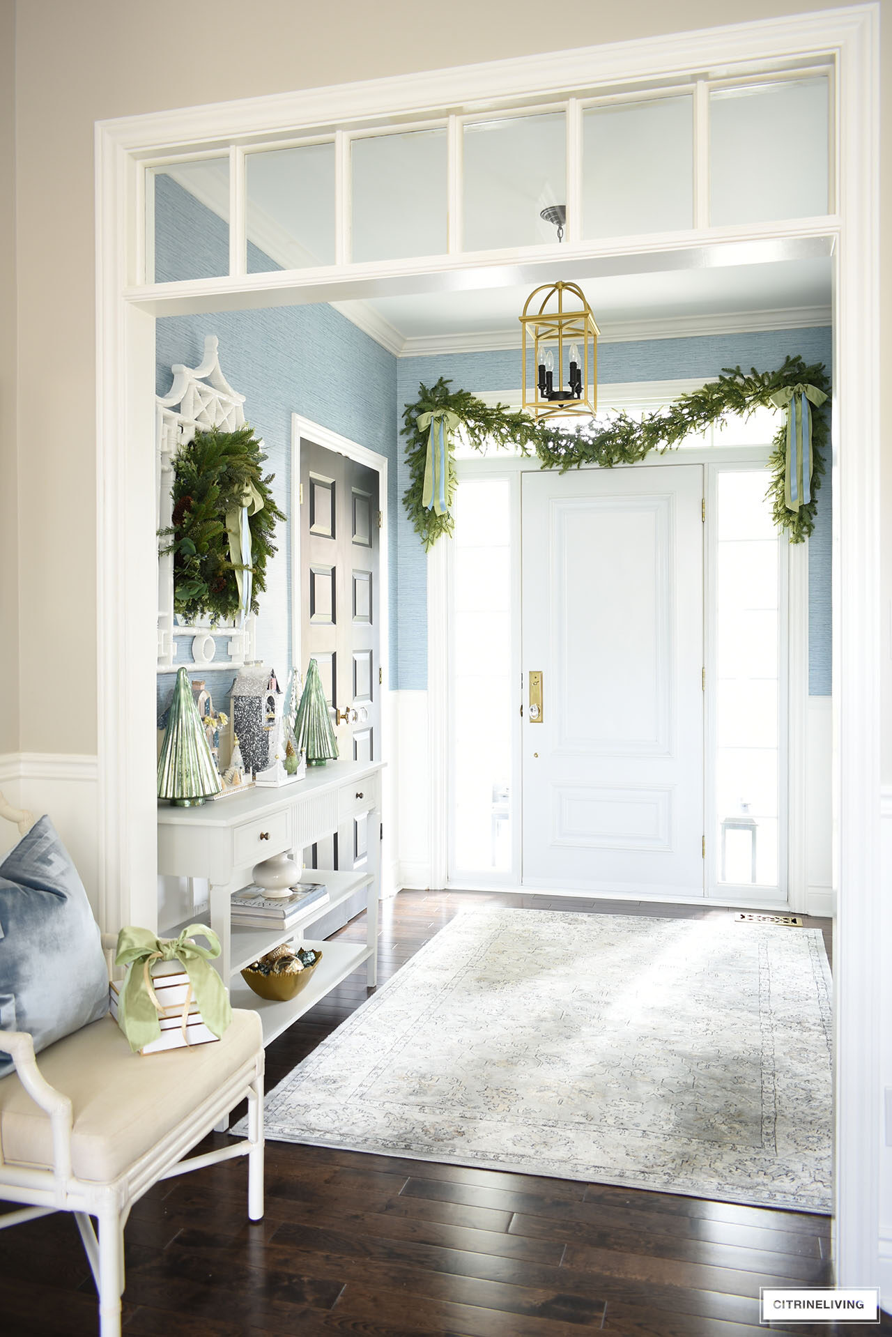 A gorgeous Christmas entryway styled with lush greenery - a garland swagged over the front door, and a wreath hung on a mirror over a console table styled with mercury glass Christmas trees and Christmas houses.