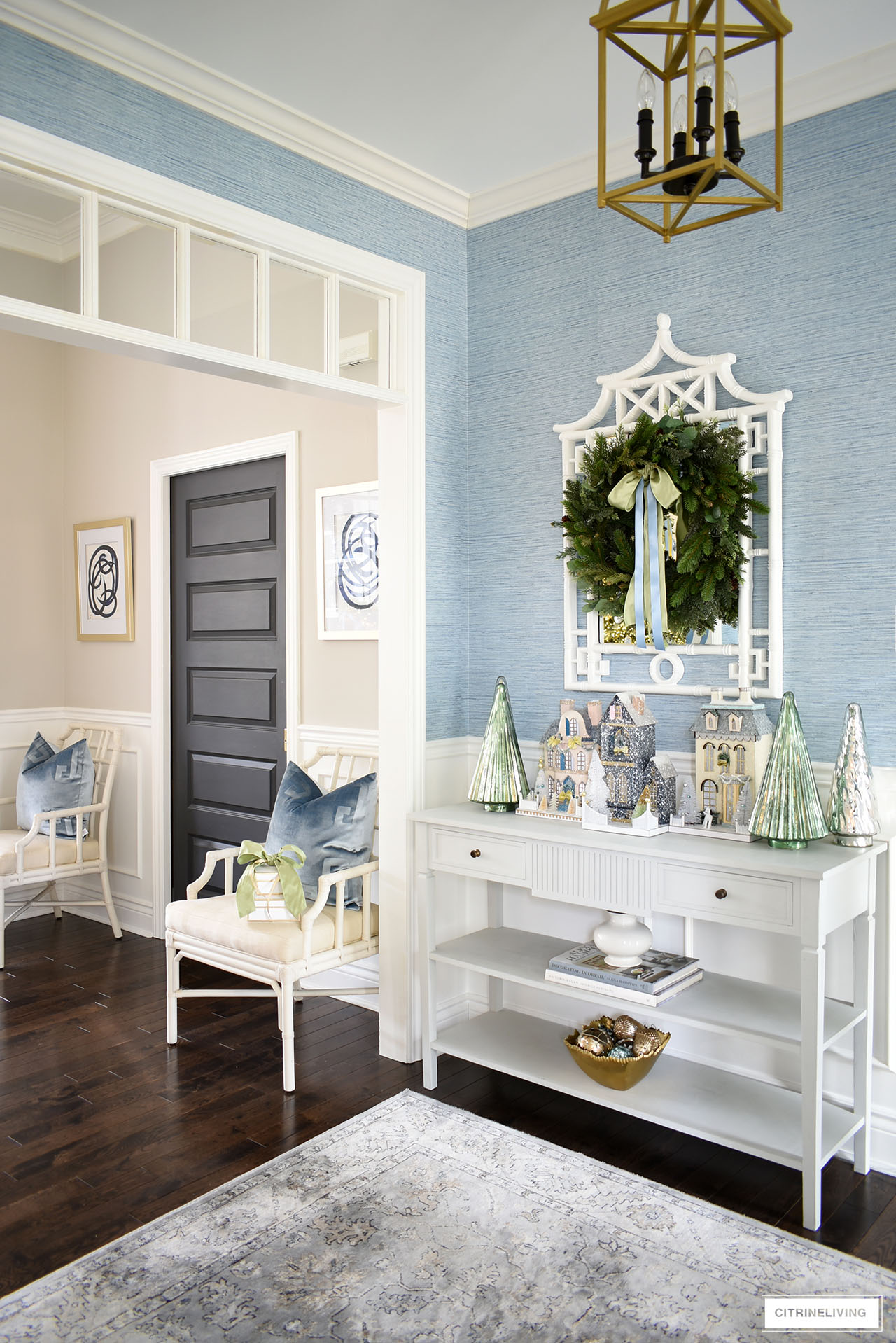 An elegant Christmas entryway decorated with a beautiful green wreath hung on a white pagoda mirror, which hangs above a console table decorated with Christmas houses and mercury glass trees.