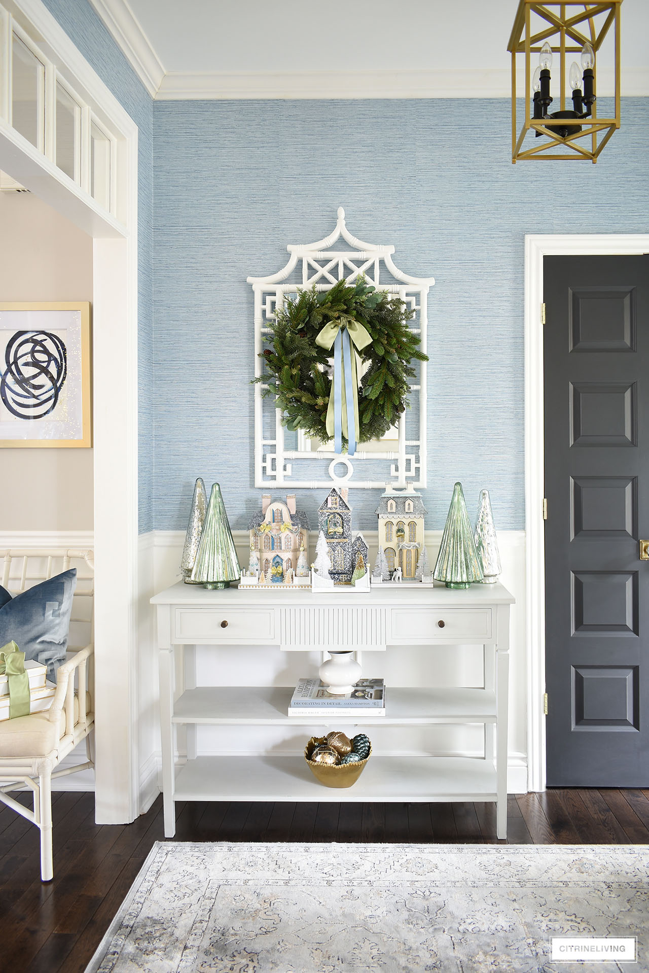 Elegant Christmas entryway decor with Christmas village houses and mercury glass trees styled on a classic console table. A lush green wreath adorned with elegant light green and light blue ribbons hangs above on a white pagoda mirror.