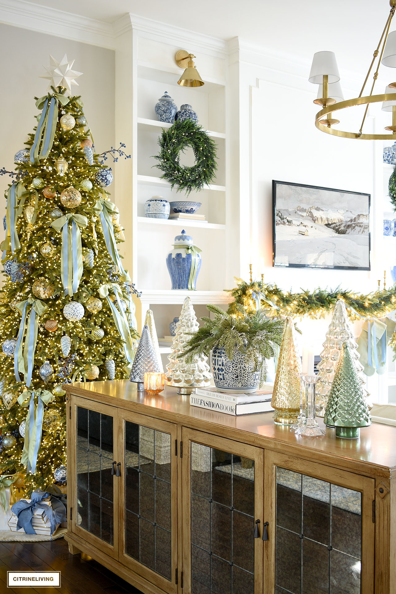 A wood and mirrored buffet decorated for Christmas with mercury glass trees in silver, blue, gold and green with a greenery arrangement in a blue and white ginger jar.