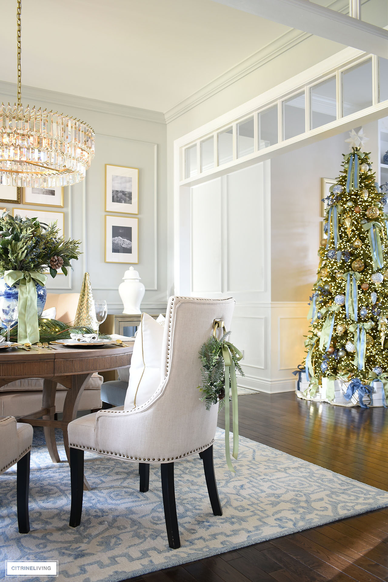 Dining room decorated for Christmas with greenery accents, blue and white chinoiserie, silver and gold table settings and a view of a Christmas tree in the next room.