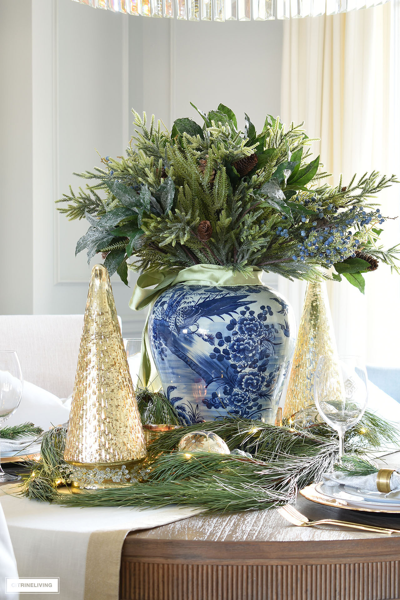 Holiday arrangement with greenery, a blue and white ginger jar and sage green ribbon tied in a bow. Mercury glass christmas trees and fresh pine branches create an elegant cetnerpiece