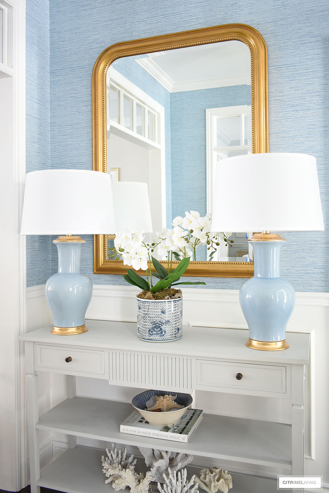 Entryway decorated for spring featuring a pair of blue lamps, large gold statement mirror with faux blue grasscloth wallpaper