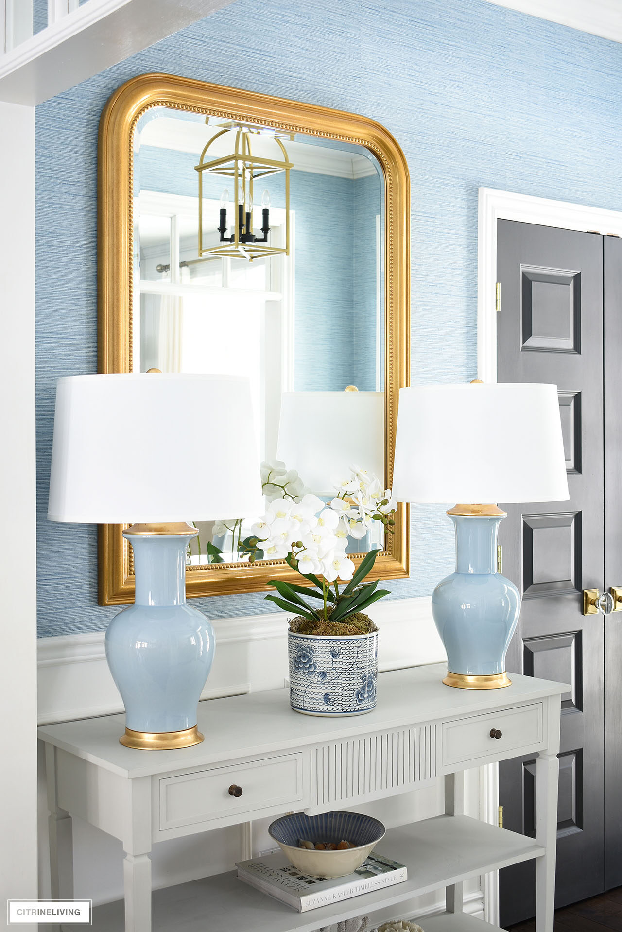 Entryway console table styled for spring with a pair of blue lamps flanking an elegant orchid arrangement styled in front of a large gold mirror