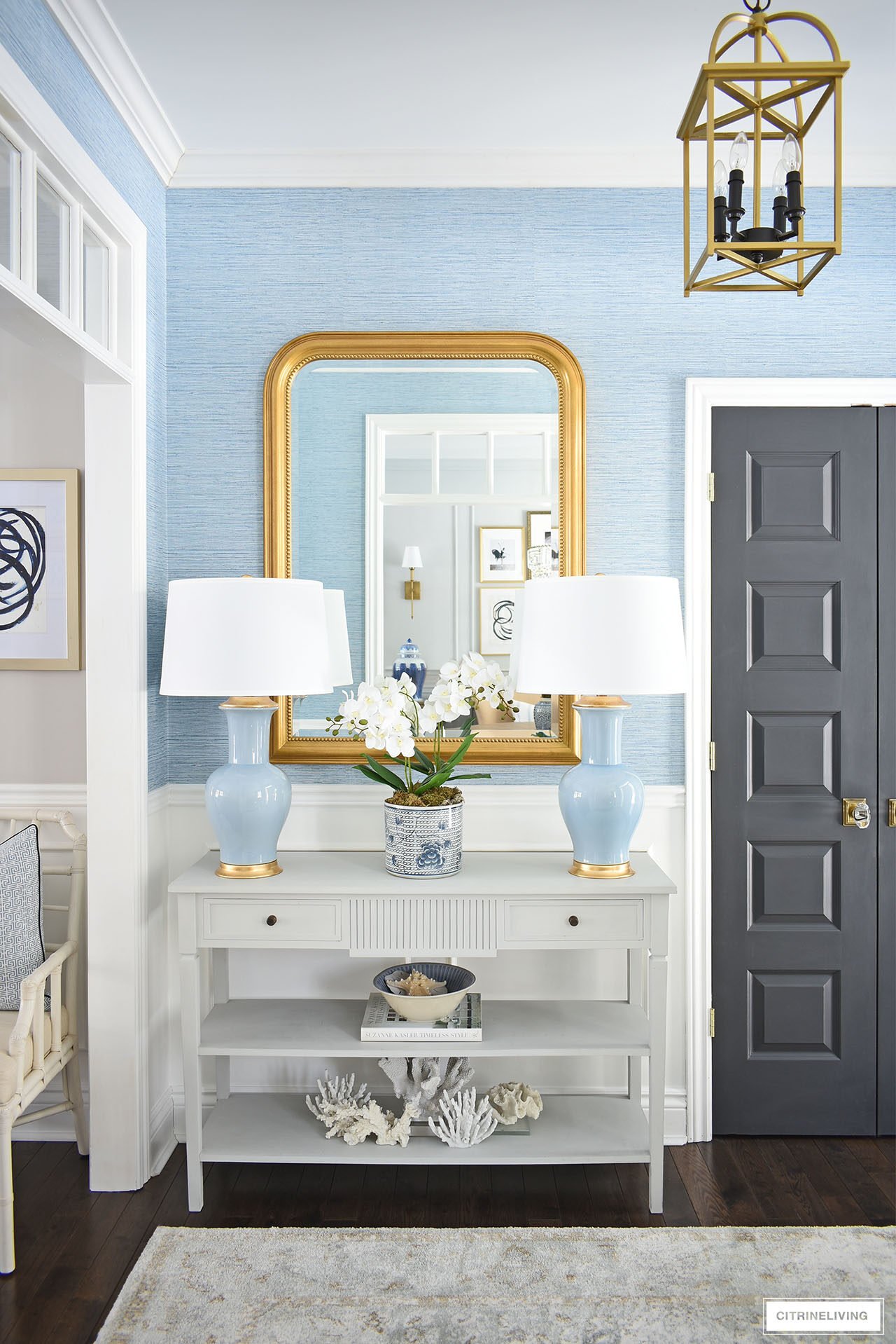 Entryway decorated for spring featuring a pair of blue lamps, large gold statement mirror and a soft muted rug, with faux blue grasscloth wallpaper