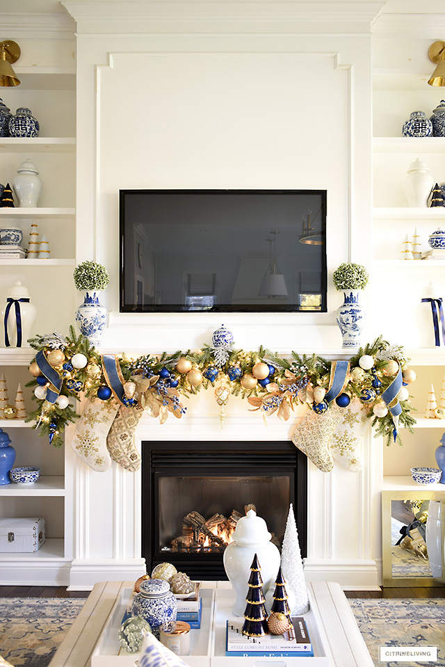 Christmas mantel decorated with faux green garlands, blue and gold ornaments and ribbon with gold and ivory beaded stockings. Blue and white chinoiserie vases with green topiary balls sit at each end of the garland on the mantel.