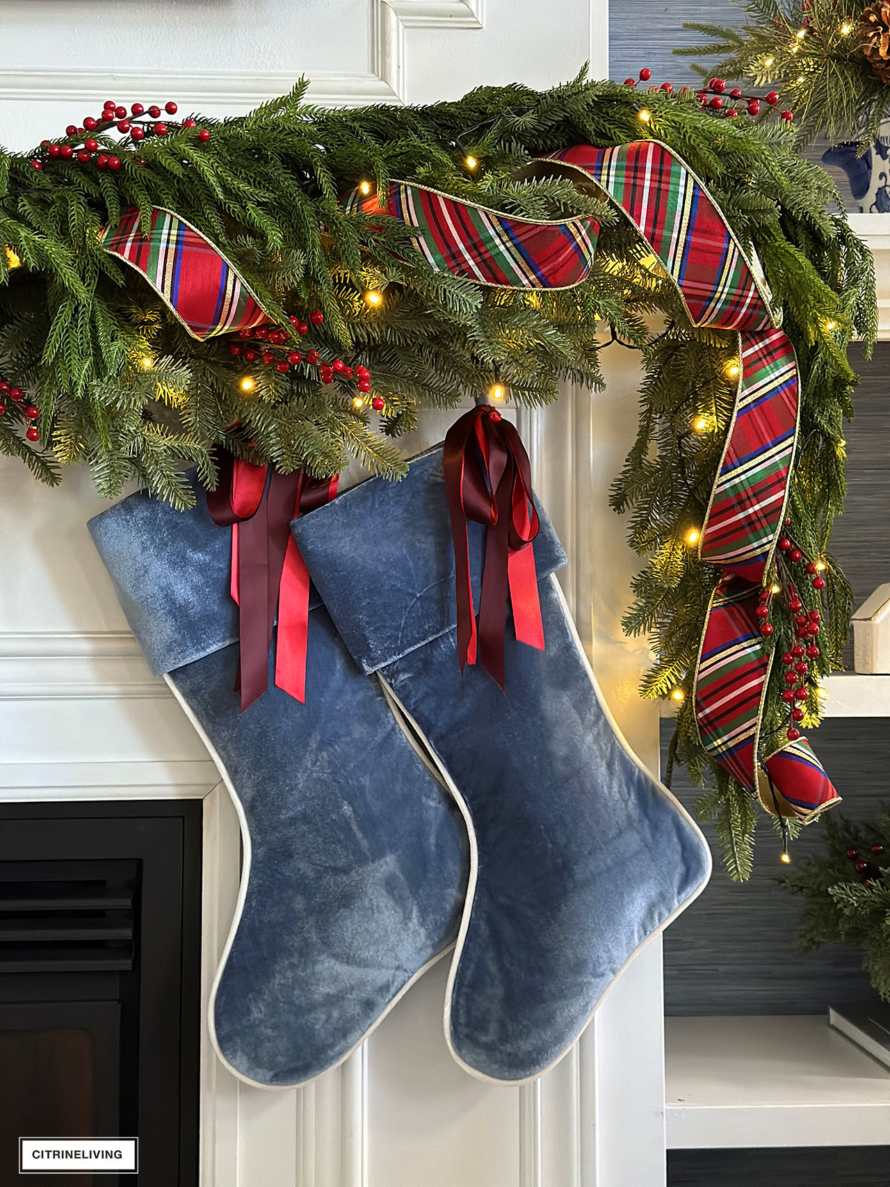 Christmas mantel with garland, red berries, red plaid ribbon and blue velvet stockings.